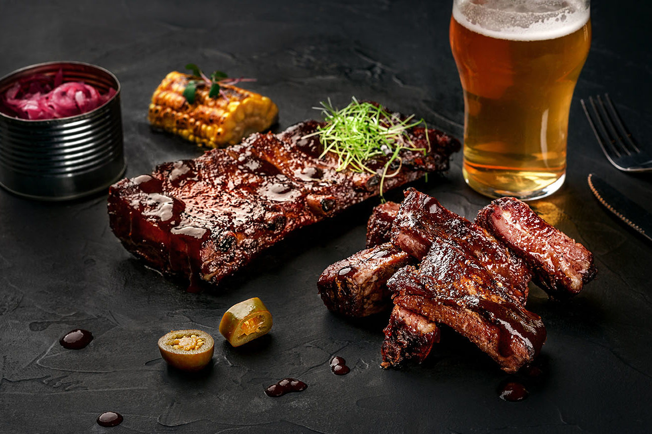 Pork ribs in barbecue sauce and a glass of beer on a black slate dish. A great snack to beer on a dark stone background. Top view with copy space